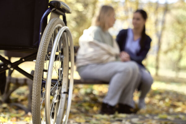 Rollstuhl mit zwei Menschen auf Bank im Hintergrund © Westend61 / gpointstudio