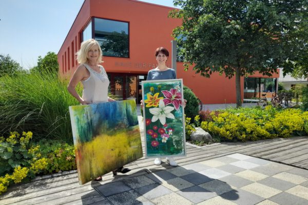 Zwei Frauen präsentieren zwei Gemälde vorm Haus der Familie © Stadt Burghausen