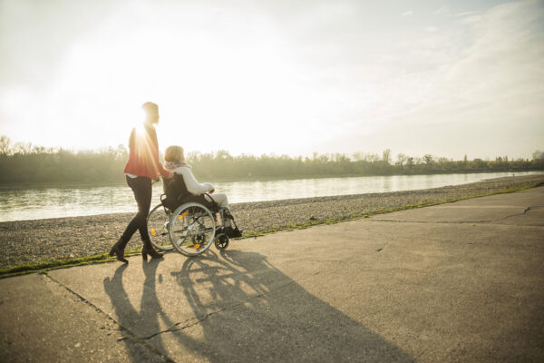 Frau schiebt andere Frau in Rollstuhl an einem Fluss entlang © Westend61 / Uwe Umstätter