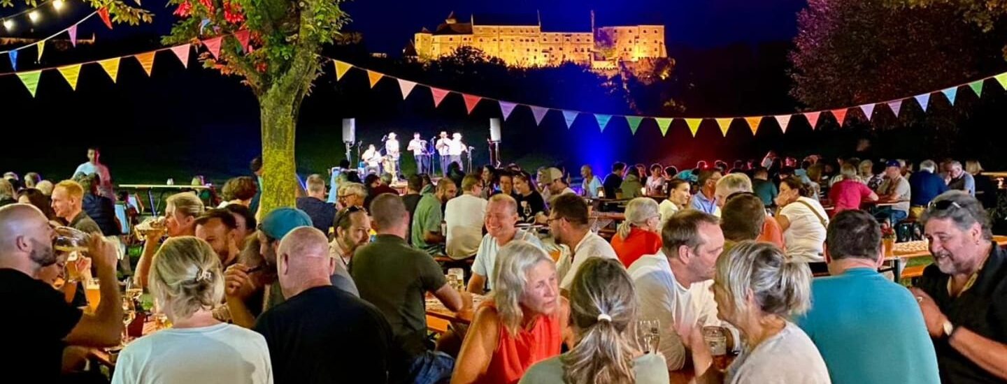 Besucher sitzen im Sommer nachts in einem Biergarten unter bunten Girlanden, im Hintergrund die beleuchtete Burghauser Burg.