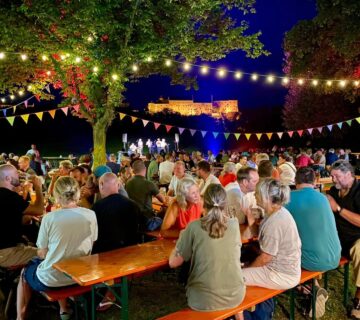 Besucher sitzen im Sommer nachts in einem Biergarten unter bunten Girlanden, im Hintergrund die beleuchtete Burghauser Burg 