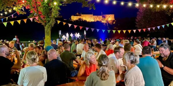 Besucher sitzen im Sommer nachts in einem Biergarten unter bunten Girlanden, im Hintergrund die beleuchtete Burghauser Burg.