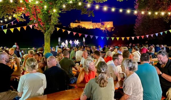 Besucher sitzen im Sommer nachts in einem Biergarten unter bunten Girlanden, im Hintergrund die beleuchtete Burghauser Burg.