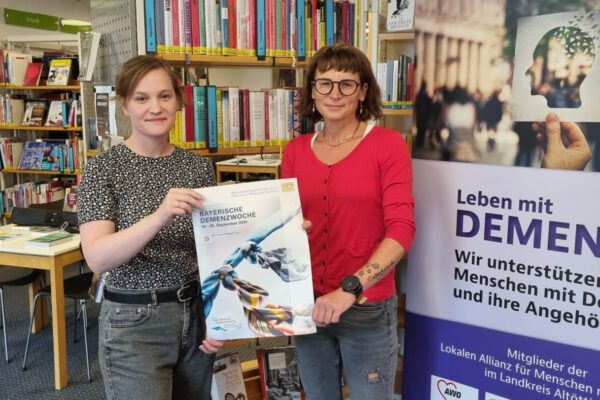 Veronika Alfranseder, Leiterin Seniorenbüro und Quartiersmanagerin der Stadt Burghausen und Christin Moll, Leiterin der Stadtbibliothek Burghausen präsentieren das Plakat zur 6. Bayerischen Demenzwoche, an der sich auch Burghausen mit einem eigenen Programm beteiligt. Fotocredit: Stadt Burghausen / köx