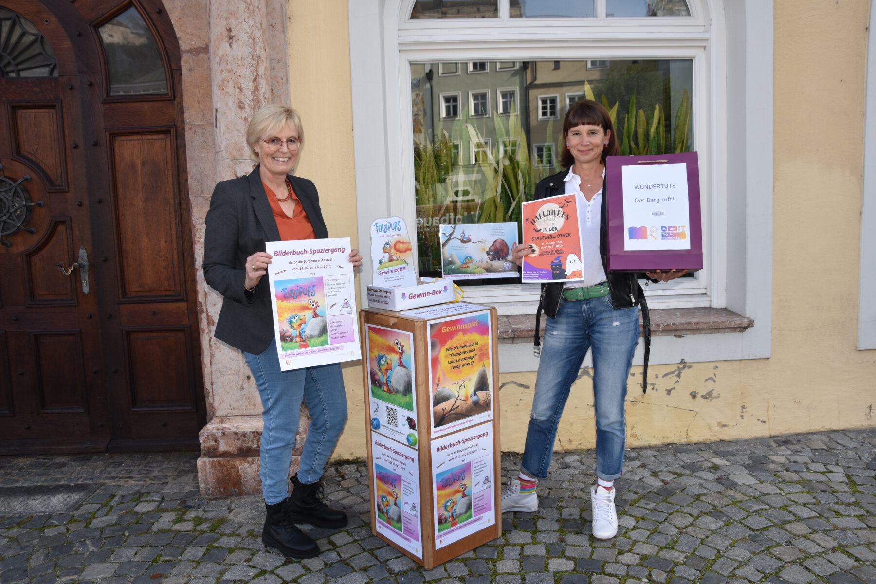 Alles ist vorbereitet: Mitarbeiterin Rosi Pfeifenthaler (l.) und Bibliotheksleiterin Christin Moll zeigen die Aktionen zum Tag der Bibliotheken am 24. Oktober, Bilderbuchspaziergang und Bücherwundertüte. Fotocredit: Stadt Burghausen / tg