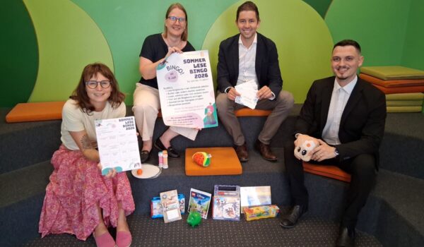 2026_07_07_Lesebingo_Stadtbücherei_Sparkasse_1_s Foto: Vier Stempel einsammeln und – Bingo! So funktioniert der Sommerlesespaß in der Stadtbibliothek. Das Team der Stadtbibliothek mit Leiterin Christin Moll und Mitarbeiterin Monika Vornehm ist bereit für den Kinderansturm, Sebastian Rauschecker und David Gergely-Komaromi von der Sparkasse Burghausen sponserten dieses Mal die Sofortgewinne. Fotocredit: Stadt Burghausen/tg
