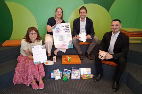 2026_07_07_Lesebingo_Stadtbücherei_Sparkasse_1_s Foto: Vier Stempel einsammeln und – Bingo! So funktioniert der Sommerlesespaß in der Stadtbibliothek. Das Team der Stadtbibliothek mit Leiterin Christin Moll und Mitarbeiterin Monika Vornehm ist bereit für den Kinderansturm, Sebastian Rauschecker und David Gergely-Komaromi von der Sparkasse Burghausen sponserten dieses Mal die Sofortgewinne. Fotocredit: Stadt Burghausen/tg