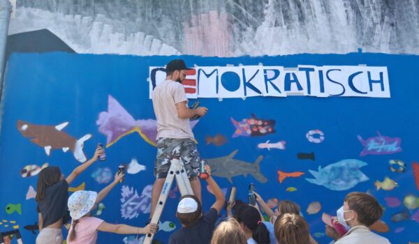 2026_07_28_Stadt_Burghausen_Graffiti_Workshop_Schülerparlament_1 Farbenrausch in der Unterwasserwelt: Graffiti-Künstler Martin Keindl leitete die Kinder bei der Umsetzung ihrer kreativen Ideen an, die Kinder reichten ihm die Spraydosen für den Schriftzug. Fotocredit: Stadt Burghausen/tg