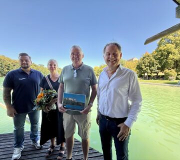 Erster Bürgermeister Florian Schneider und Bäderleiter Florian Moser begrüßten das Ehepaar Rosmarie und Richard Siegerd aus Haiming als 100 000 Badegäste  Das Ehepaar ist Stammgast und besucht den See mindestens vier Mal pro Woche  „Wir schätzen die sehr gute Wasserqualität und die natürliche Beschattung  Das Flair ist einzigartig und wenn wir am Vormittag da sind ist es auch noch schön ruhig“, so die Siegerds  Als kleine Aufmerksamkeit überreicht das Stadtoberhaupt Blumen und eine Saisonkarte für das nächste Jahr 
